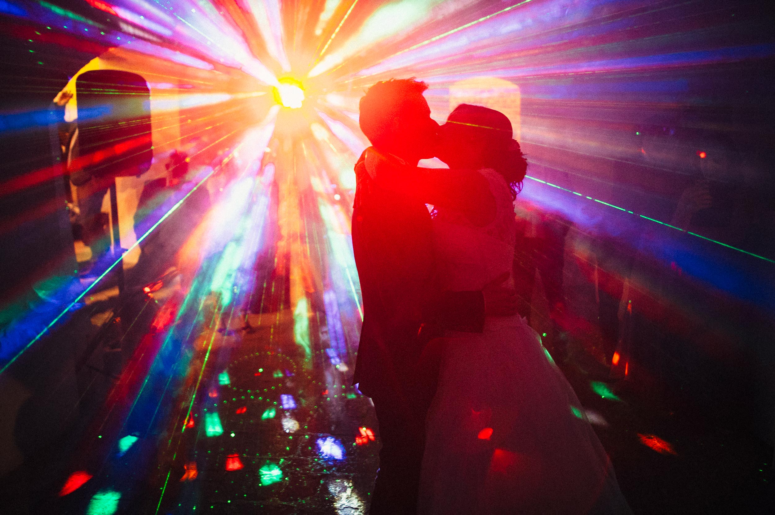 dj lights color flare strikes on the dancefloor while bride and groom kiss during the first dance at wedding