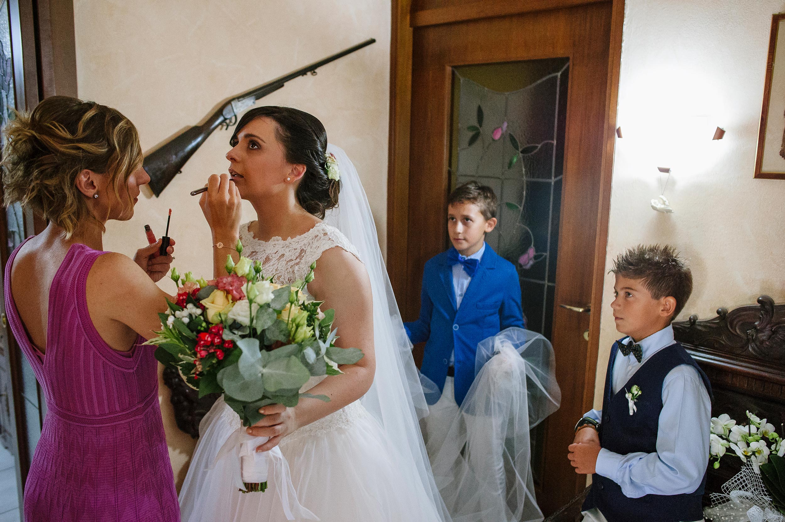 bride doing last make up retouches rifle on the wall kids holding the veil